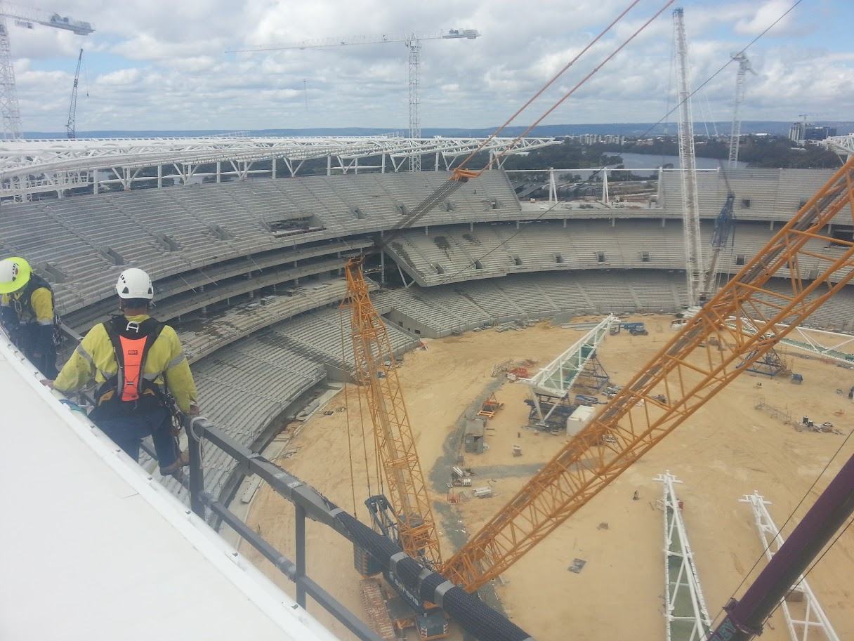 Rope access technician in hi-vis and helmet at the roof edge of a stadium build, with tower cranes and the arena floor below