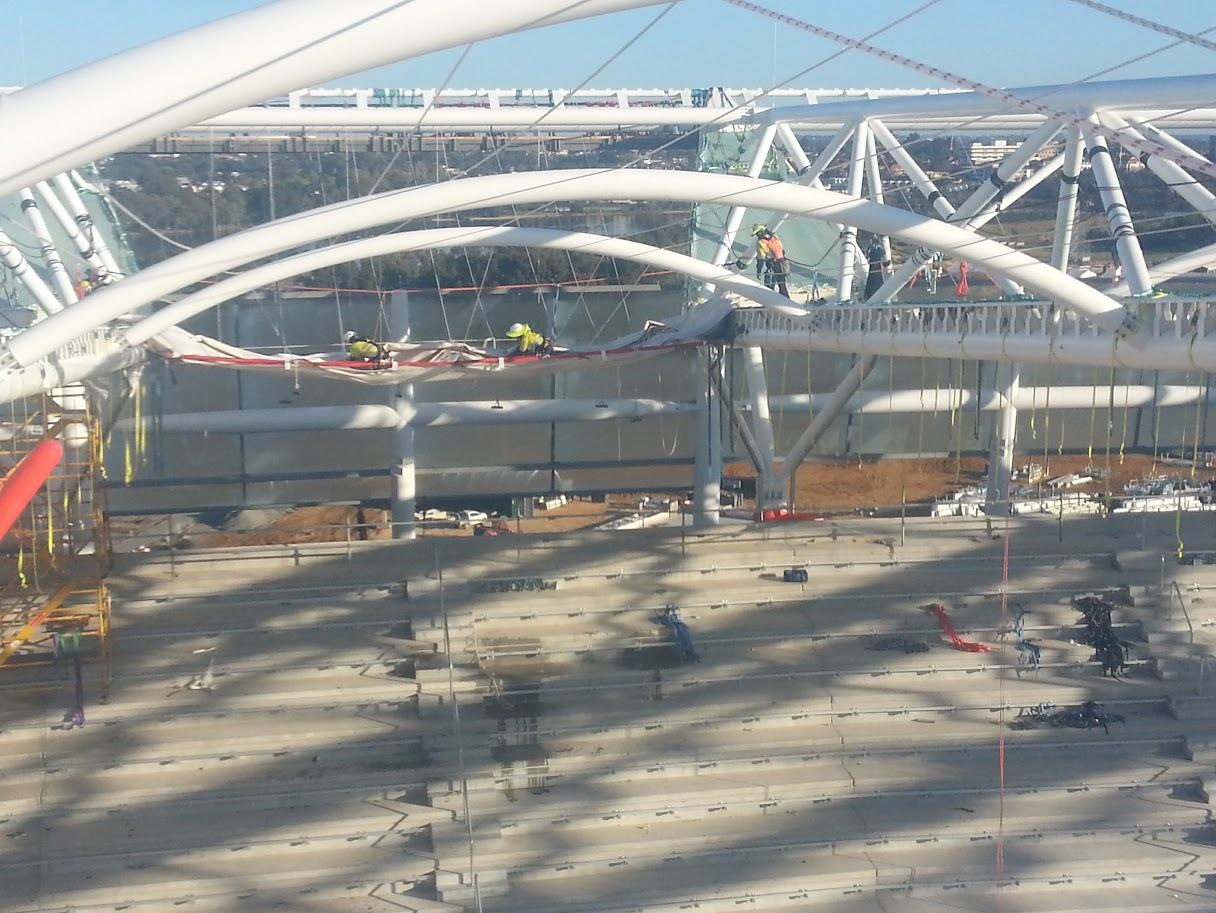 White arched roof trusses spanning a stadium under construction, with rope access workers and rigging along the steelwork