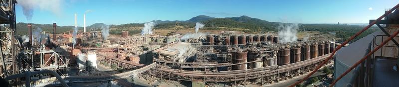 Panorama of a processing plant with pipework and structures, and green hills in the distance