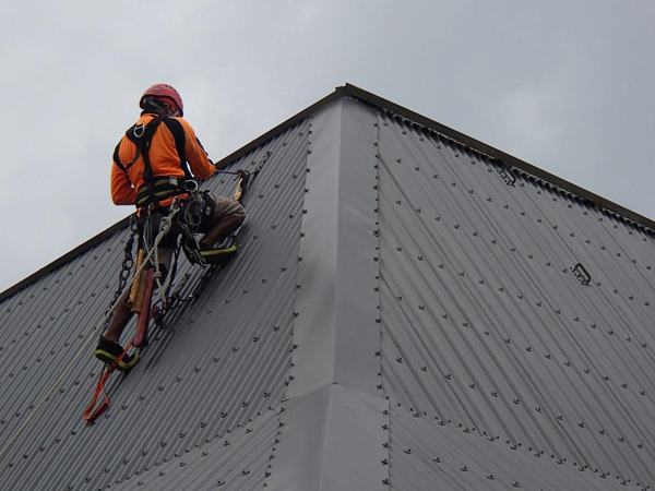 Rope access technician in orange climbing on rope up a steel-clad roof structure