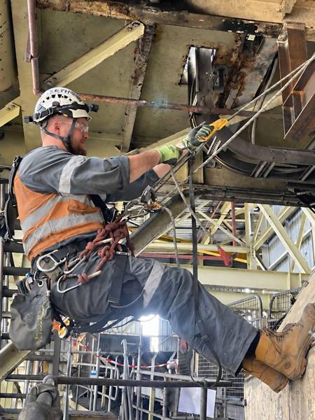 Rope access technician in orange hi-vis and helmet positioned on rope among industrial steelwork
