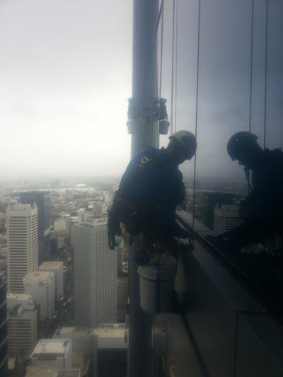 Rope access technician on a glass tower façade with a cleaning bucket, high above the city