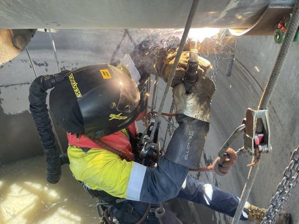 Technician in a welding helmet and hi-vis carrying out hot work while suspended on rope inside a tank